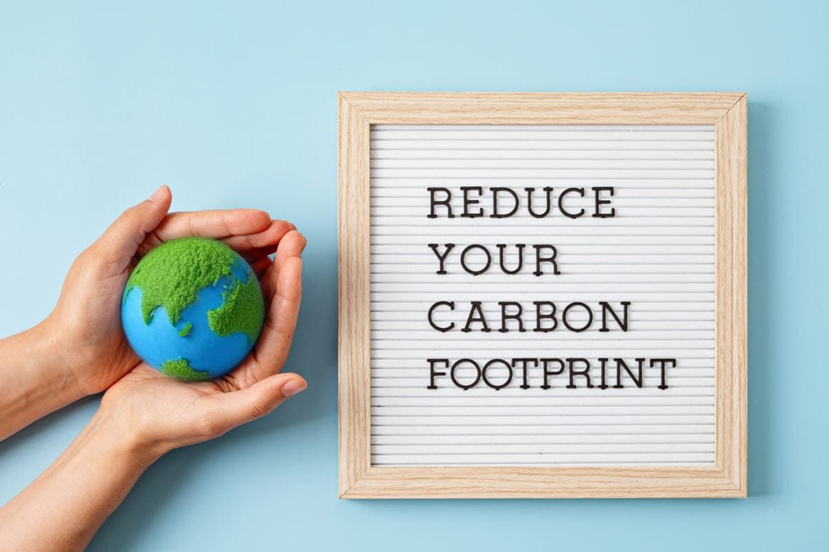 Mani sorreggono un modellino della Terra accanto a un pannello con la scritta "Reduce Your Carbon Footprint", simbolo della Carbon Footprint aziendale.